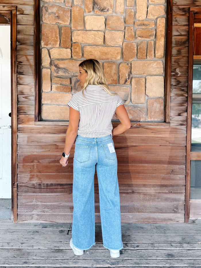 A woman stands with her back to the camera wearing a white and brown striped short-sleeve shirt and light blue wide-leg jeans, posing in front of a rustic wooden and stone wall.