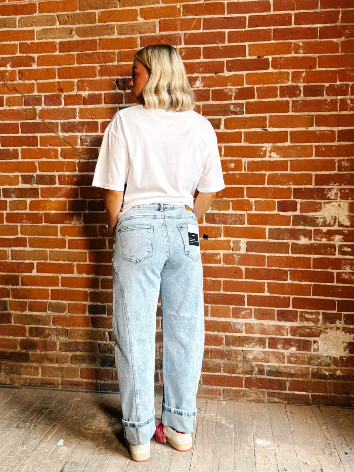 A person standing with their back to the camera, wearing light-wash blue jeans, a white t-shirt, and pink sneakers, set against a red-brown brick wall background.