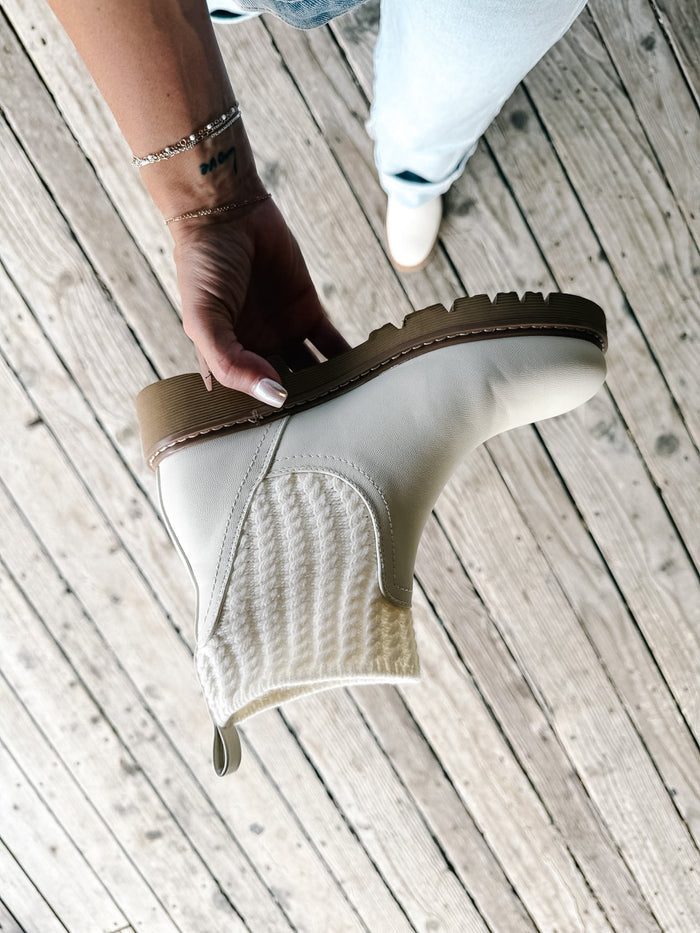 A hand holds up a cream-colored ankle boot with a textured knit side panel and a brown sole, set against a background of light-colored wooden planks with a person's legs and jeans visible.