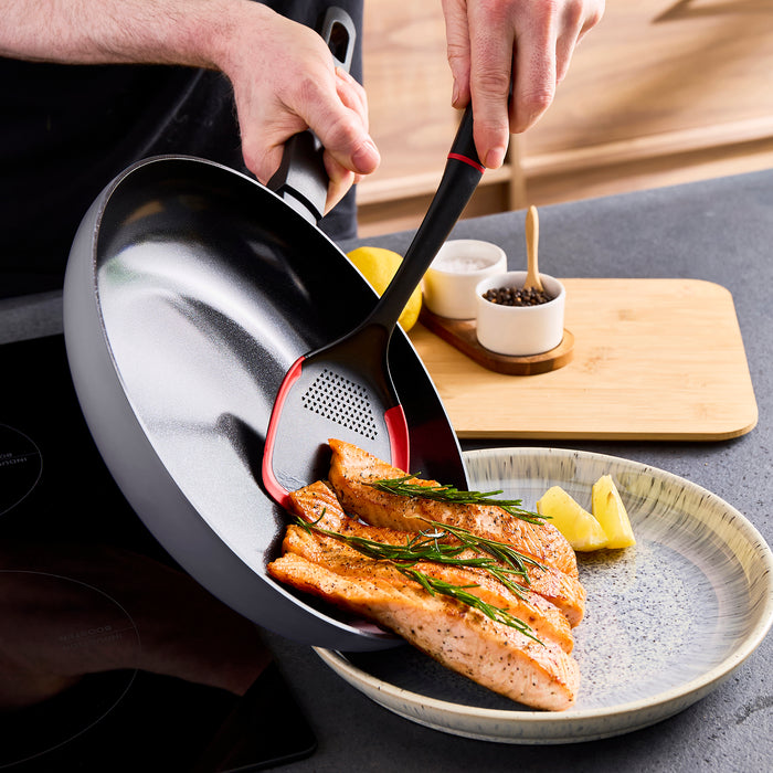 Person using a frying pan to cook salmon on a plate with lemon and herbs.