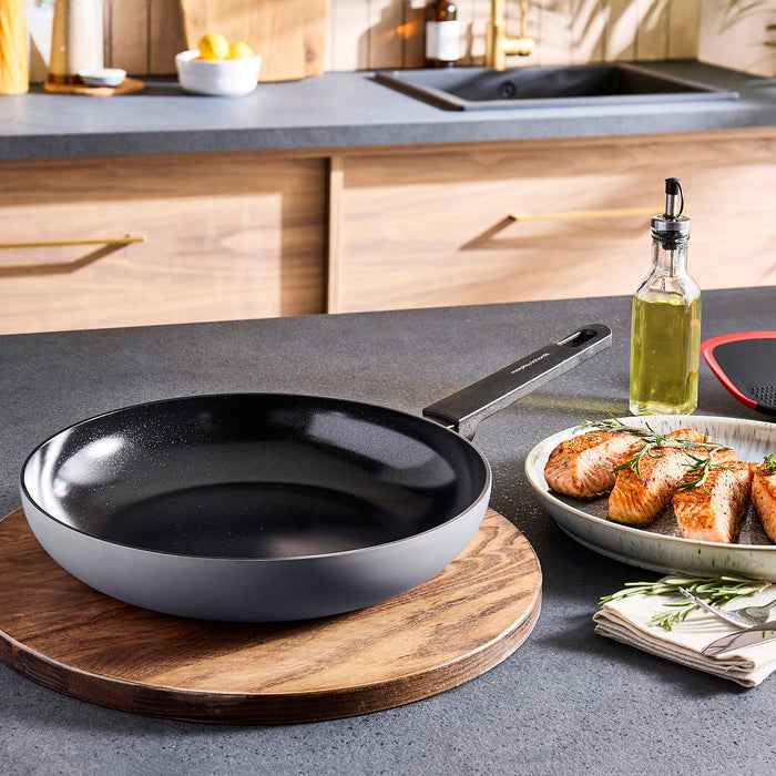 Black frying pan on a kitchen counter with a plate of food and bottle of oil in the background.