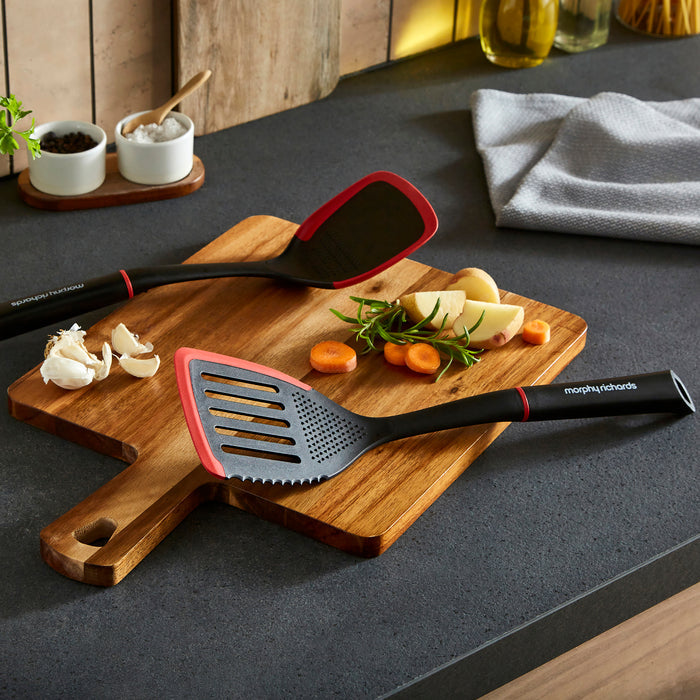 Black and red spatula on a wooden cutting board with vegetables on a kitchen counter.