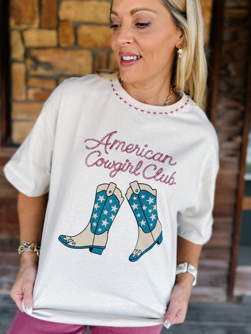 A woman wearing a white graphic t-shirt with 'American Cowgirl Club' in pink script and a pair of blue and beige cowboy boots with white stars. The background shows a rustic stone wall and wooden door.
