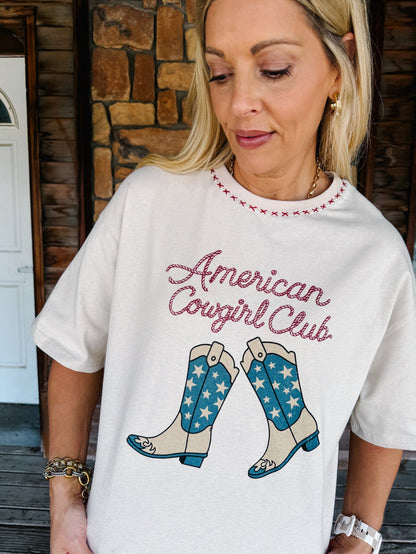 A woman wearing a white graphic t-shirt with 'American Cowgirl Club' in pink script and a pair of blue-and-white star-patterned cowboy boots, posed in front of a rustic stone wall.