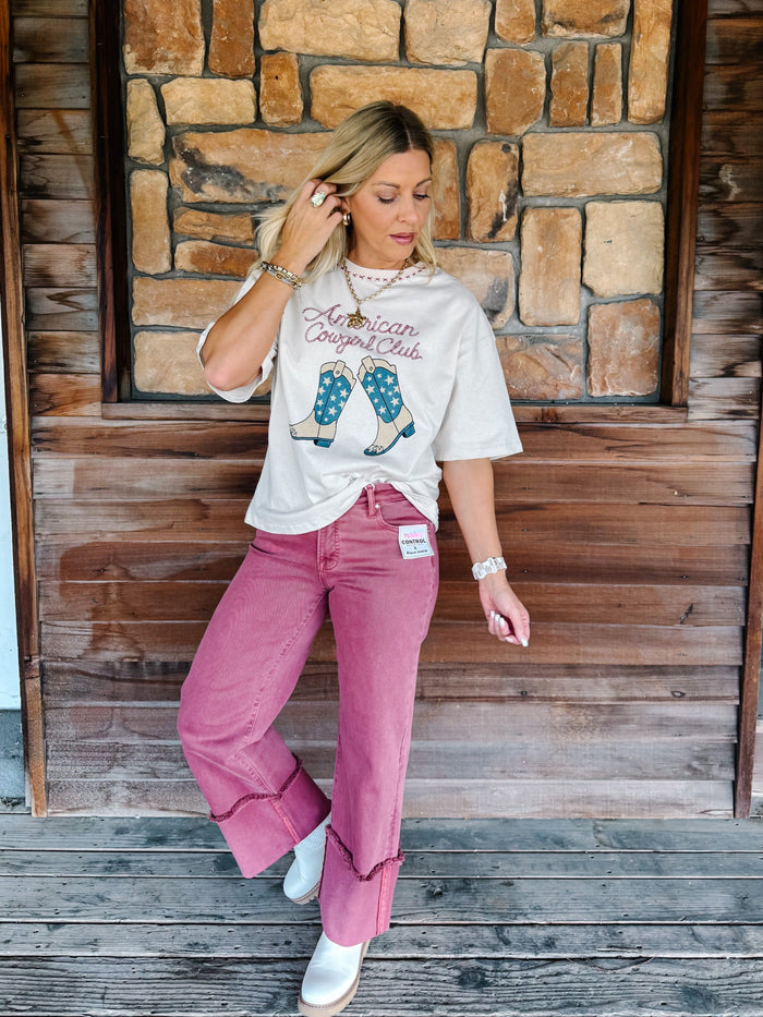 A woman posing in front of a rustic wooden and stone wall, wearing a cream graphic t-shirt with 'American Originals' text and blue polka-dot boot graphics, paired with dusty rose wide-leg jeans and white sneakers.
