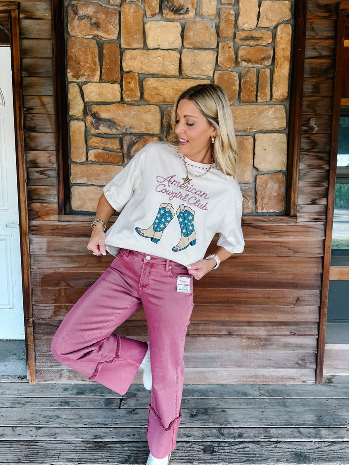 A woman posing on a rustic wooden porch in front of a stone wall, wearing a cream graphic t-shirt that reads 'American Cowboy Club' with cowboy boot illustrations, and dusty rose wide-leg jeans.
