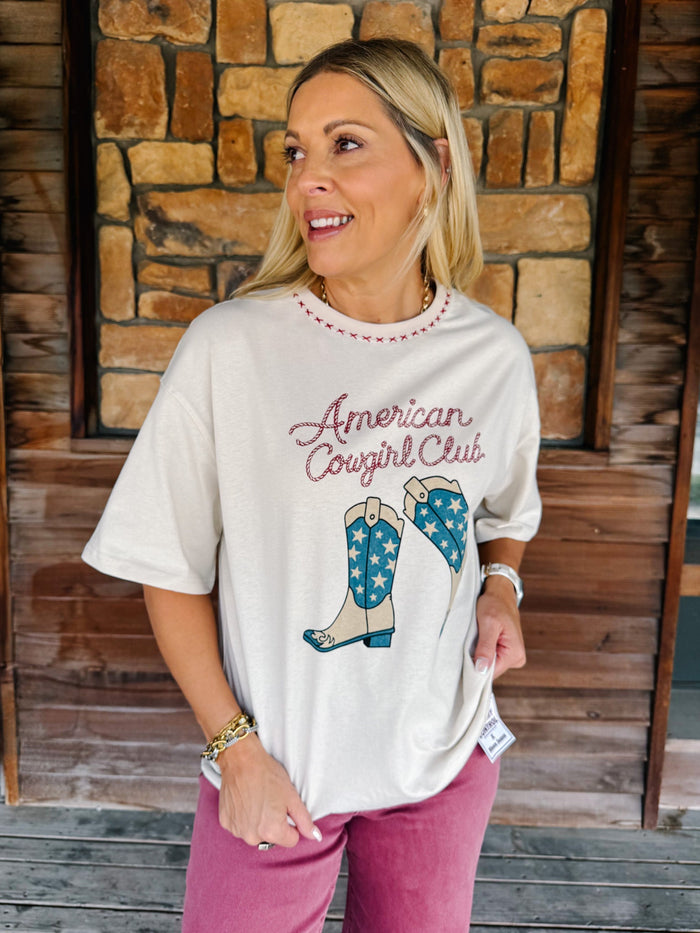 A woman wearing a cream-colored graphic t-shirt with 'American Cowgirl Club' and cowboy boots printed on the front, paired with pink pants, standing in front of a rustic stone and wood wall.