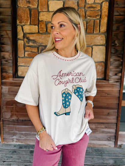 A woman wearing a cream-colored graphic t-shirt with 'American Cowgirl Club' and cowboy boots printed on the front, paired with pink pants, standing in front of a rustic stone and wood wall.