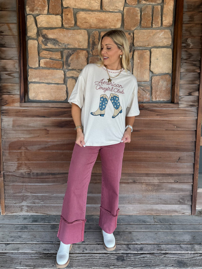 A woman stands on a wooden porch in front of a stone wall, wearing a cream graphic t-shirt that reads 'American Cowboy Club' with an illustration of cowboy boots, dusty rose wide-leg pants, and white boots.