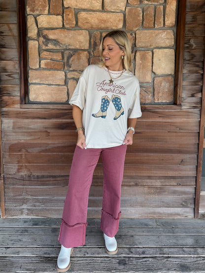 A woman stands on a wooden porch in front of a stone wall, wearing a cream graphic t-shirt that reads 'American Cowboy Club' with an illustration of cowboy boots, dusty rose wide-leg pants, and white boots.
