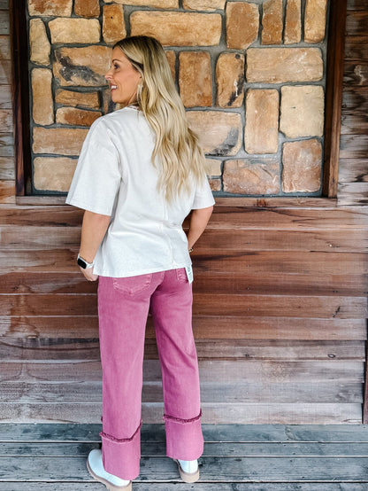 A woman stands with her back to the camera wearing a white short-sleeve top and pink wide-leg jeans with rolled cuffs, posed in front of a rustic stone and wood wall.