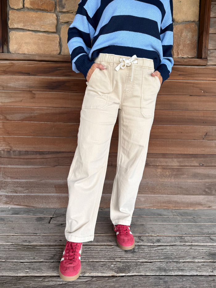 A person wearing beige drawstring pants, a blue and navy striped sweater, and pink sneakers, standing against a rustic wooden wall and stone floor.