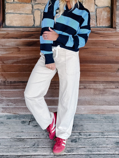 A person wearing a blue and navy striped knit sweater, cream wide-leg sweatpants, and pink sneakers, standing against a rustic wooden wall and stone floor.