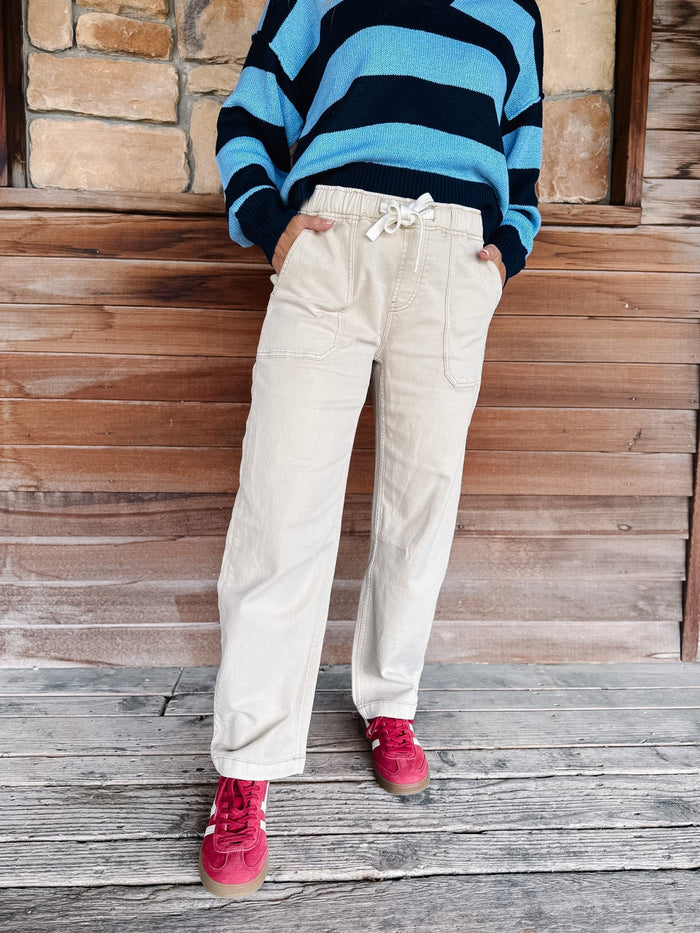 A person wearing cream-colored wide-leg pants with a white drawstring waist, paired with a blue and black striped sweater and pink sneakers, standing against a rustic wooden wall.