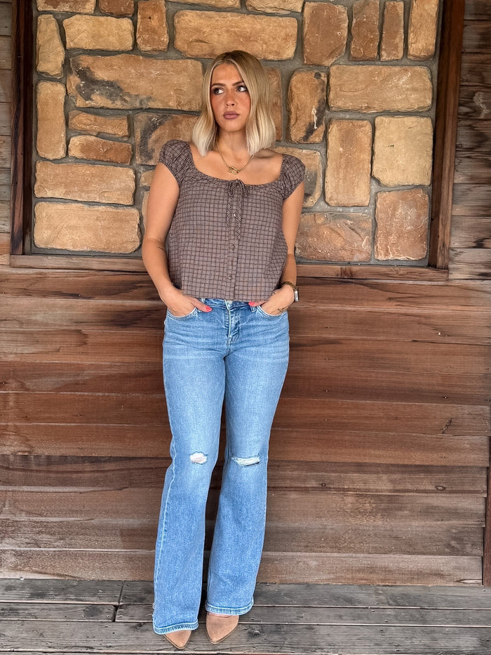 A woman stands against a rustic stone and wood wall wearing a brown and black gingham patterned crop top with cap sleeves, paired with light blue distressed flare jeans and beige heels.