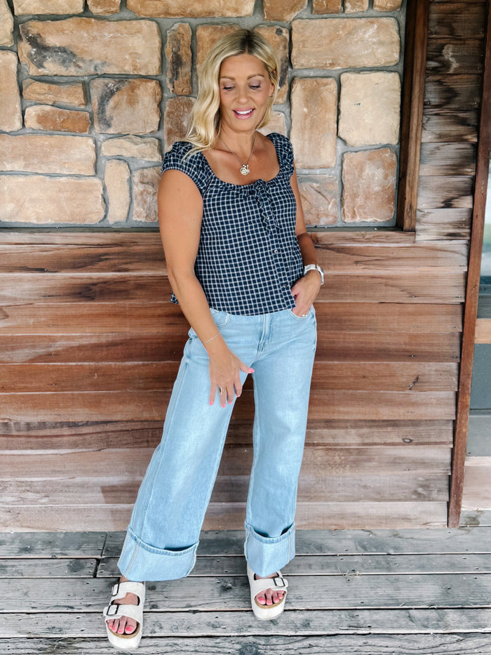A woman stands on a wooden deck in front of a stone wall, wearing a navy and white gingham sleeveless top, light blue wide-leg jeans, and white platform sandals.