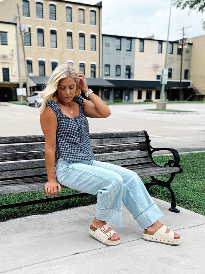 A woman sits on a wooden park bench wearing a black and white gingham sleeveless top, light blue wide-leg jeans, and beige slide sandals, with an urban street scene in the background.