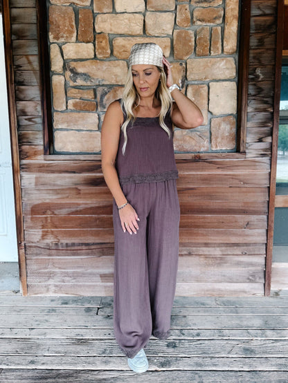A woman stands on a rustic wooden porch in front of a stone wall, wearing a sleeveless mauve-purple jumpsuit with wide legs and a textured knit head covering.