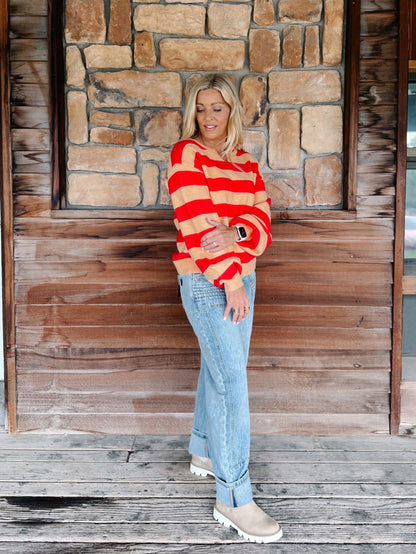 A woman stands on a rustic wooden porch in front of a stone wall, wearing a red and beige striped knit sweater, light blue jeans, and beige boots.