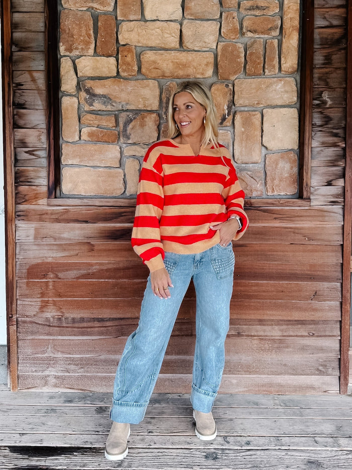 A woman stands on a rustic wooden porch in front of a stone wall, wearing a red and beige striped long-sleeve knit sweater, light-wash blue jeans, and tan boots.