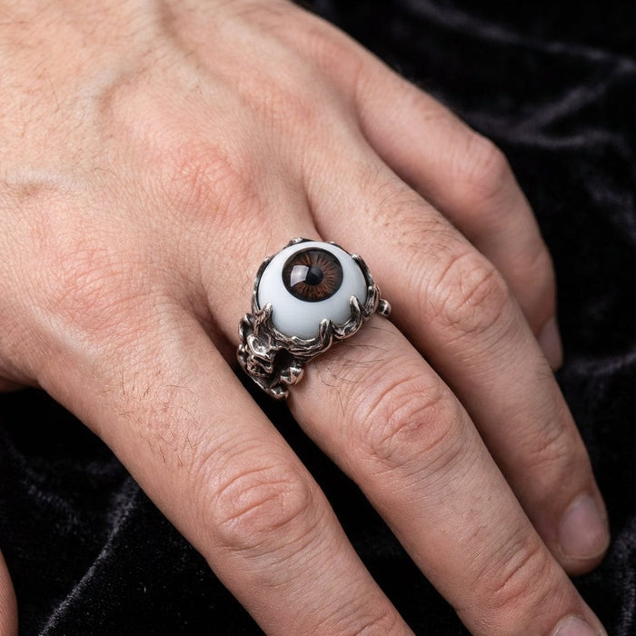 Sterling silver gothic ring featuring a realistic brown eyeball centerpiece held by ornate devil skull claws, worn on a hand against a luxurious black velvet background