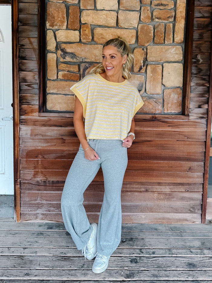 A person wearing light heather gray drawstring flare sweatpants, a yellow and heather oatmeal striped top, and white sneakers, standing against a rustic wooden wall and deck.