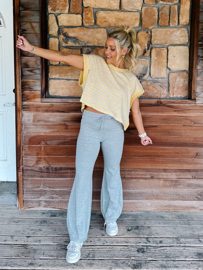 A person wearing light heather gray drawstring flare sweatpants, a yellow and heather oatmeal striped top, and white sneakers, standing against a rustic wooden wall and deck.