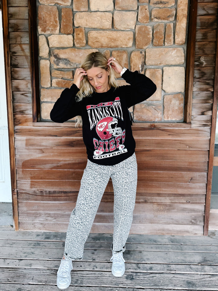 A woman stands on a wooden deck in front of a stone wall, wearing a black crewneck sweatshirt with a red and white 'Lamar Hunt Racing' graphic, patterned gray pants, and white sneakers.