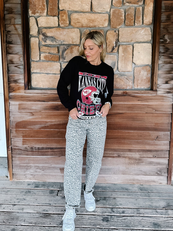 A woman stands on a wooden deck in front of a stone wall, wearing a black long-sleeve graphic sweatshirt with a red and white 'Kansas City Chiefs' football helmet design, gray patterned sweatpants, and white sneakers.