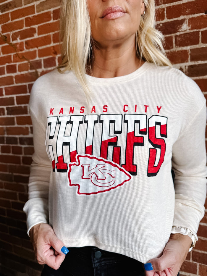 A woman wearing a cream ribbed long-sleeve crewneck sweatshirt with a large Kansas City Chiefs graphic on the front, standing against a red brick wall.