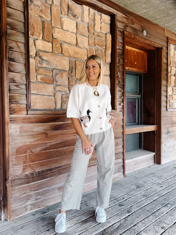A woman stands on a rustic wooden porch in front of a stone wall, wearing a cream short-sleeve button-front cardigan with duck embroidery, a patterned knit top, and white sneakers.