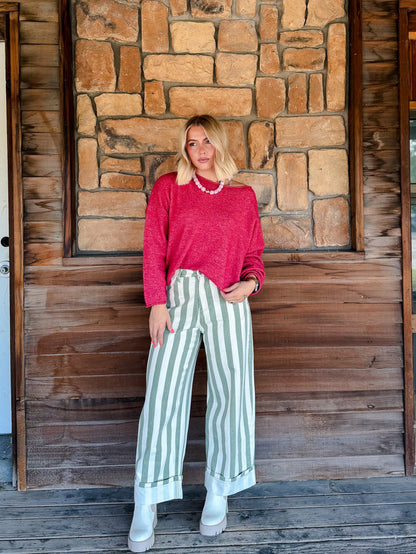 A woman stands on a wooden porch in front of a stone wall, wearing a bright pink knit sweater, light green and white vertically striped wide-leg pants, and white boots.