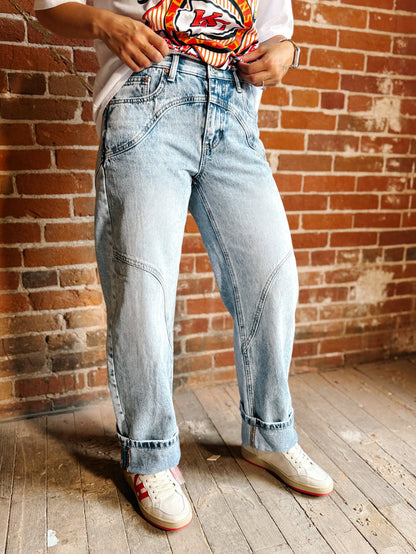 A person wearing light-wash light blue denim jeans with a relaxed straight-to-wide leg fit, cuffed hems, and contrast stitching, paired with a white t-shirt and white sneakers, standing in front of a brick wall.