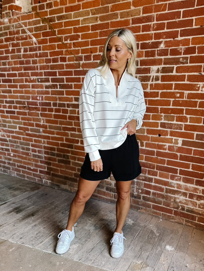 A woman posing in front of a red brick wall, wearing a white long-sleeve quarter-zip pullover with thin black horizontal stripes, black shorts, and white sneakers.