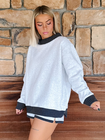 A person wearing light heather gray drawstring shorts with navy-and-white polka dot trim on the side pocket and hem, paired with a matching oversized sweatshirt. The photo is cropped from the chest down, showing the shorts and part of the sweatshirt in a real indoor setting with wood flooring and a stone wall.