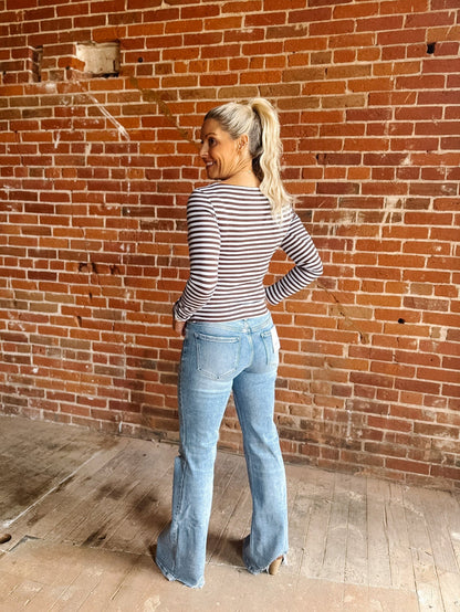 A woman wearing a brown and light blue horizontally striped long-sleeve boat-neck top, paired with blue jeans, standing against a red brick wall.