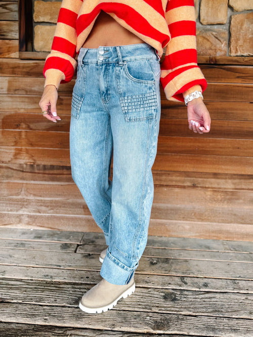 A person wearing light-wash blue jeans with decorative square stitching on the front pockets, paired with a red and beige striped cropped sweater and beige boots, standing on a wooden deck in front of a rustic stone and wood wall.
