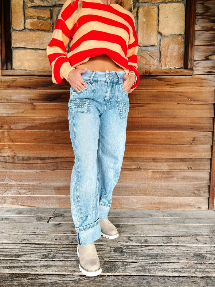 A person wearing a red and beige striped cropped sweater, light-wash blue jeans, and beige boots, standing on a wooden deck in front of a rustic stone and wood wall.