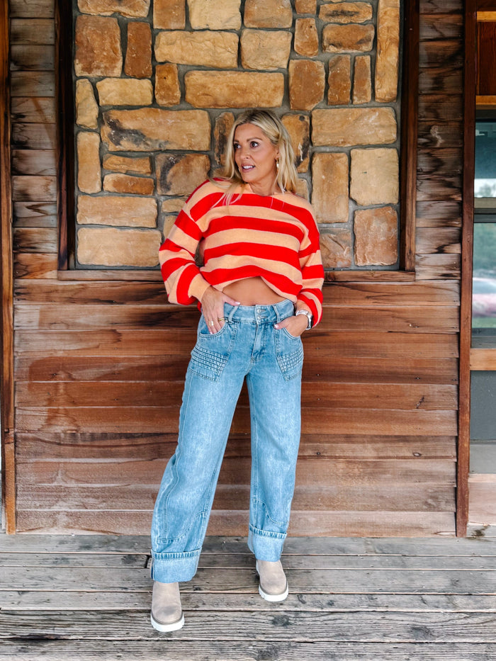 A woman stands on a rustic wooden porch in front of a stone wall, wearing a cropped red and orange striped knit sweater, light blue high-waisted jeans with decorative stitching, and gray slip-on shoes.