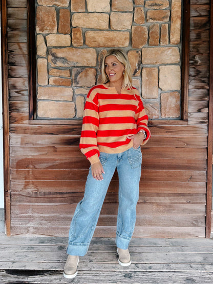 A woman stands on a wooden porch in front of a stone wall, wearing a red and beige striped knit sweater, light blue jeans, and tan boots.