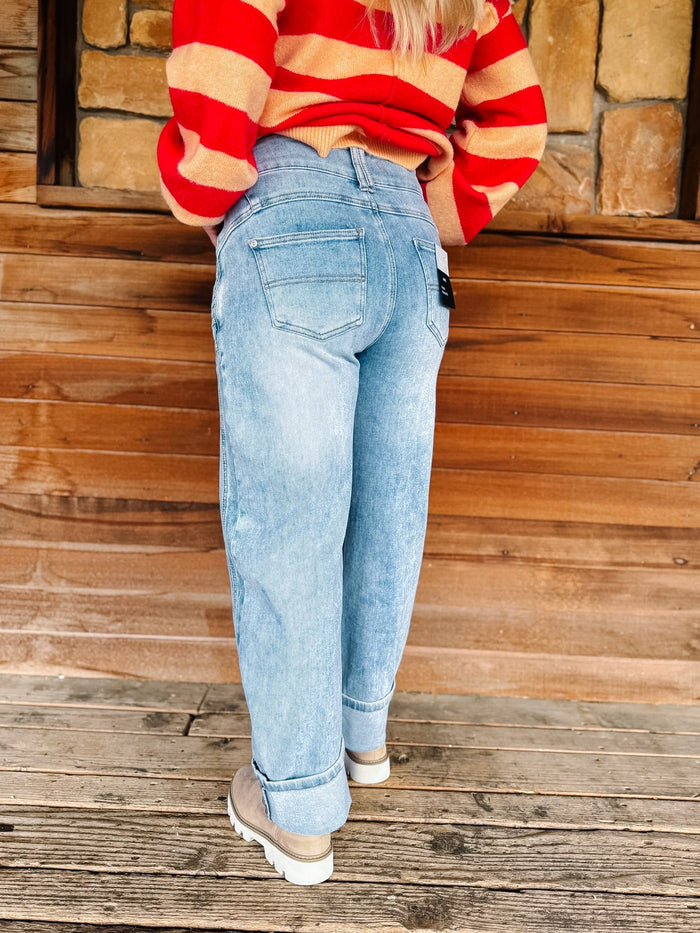 A person shown from the back wearing light-wash blue jeans, a red and beige striped sweater, and beige shoes, standing on a wooden deck in front of a stone wall.