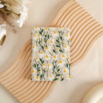 A white vegan leather Kindle case with a floral pattern, displayed on a wooden surface with a Kindle and a cup.