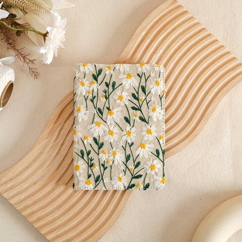 A white vegan leather Kindle case with a floral pattern, displayed on a wooden surface with a Kindle and a cup.