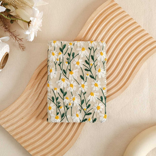 A white vegan leather Kindle case with a floral pattern, displayed on a wooden surface with a Kindle and a cup.