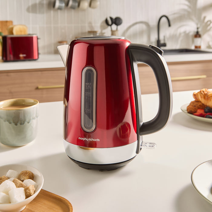 Red Morphy Richards jug kettle on a kitchen counter with a cup of coffee and breakfast items.