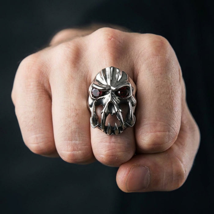 A close-up of a masculine clenched fist wearing a heavy silver gothic devil ring featuring prominent fangs and deep red gemstone eyes, set against a dark background.
