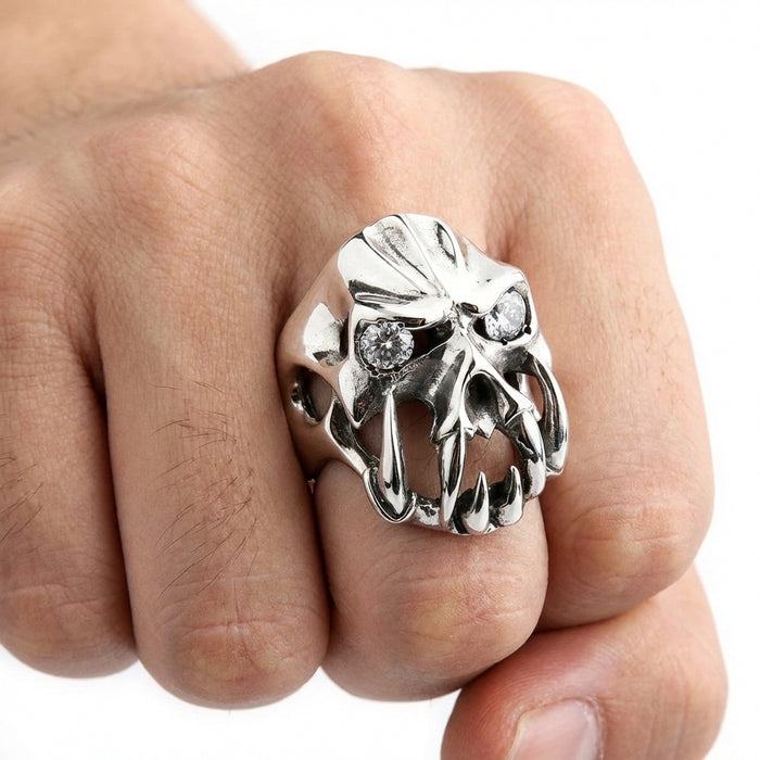 Close-up of a man's fist wearing a chunky silver gothic devil ring featuring prominent fangs and clear gemstone eyes, set against a plain white background.