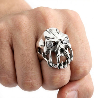 Close-up of a man's fist wearing a chunky silver gothic devil ring featuring prominent fangs and clear gemstone eyes, set against a plain white background.