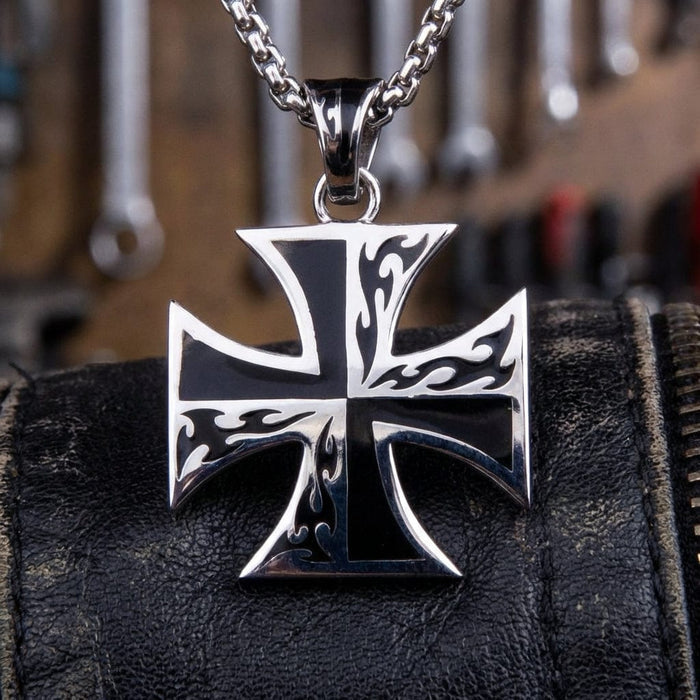Sterling silver Iron Cross pendant necklace with tribal flame design on black enamel, displayed on black leather with workshop tools in the background