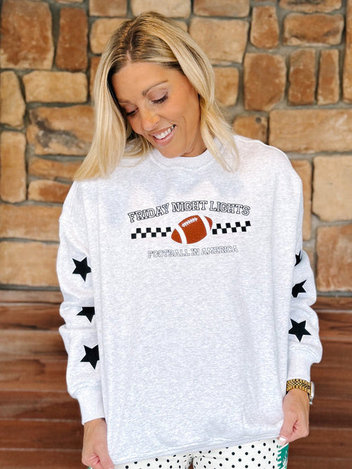 A woman wearing a white heathered long-sleeve sweatshirt with a 'Friday Night Lights Football Night in America' graphic on the front and black stars on the sleeves, standing in front of a stone wall.
